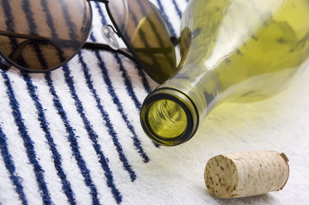 Neck of wine bottle flanked by sunglasses and cork on beach towel